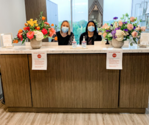 Welcoming Front desk at Memorial Hearing in Houston, TX. Two woman sit behind a counter, both wearing face masks and two big colorful bouquets on either side of them. 